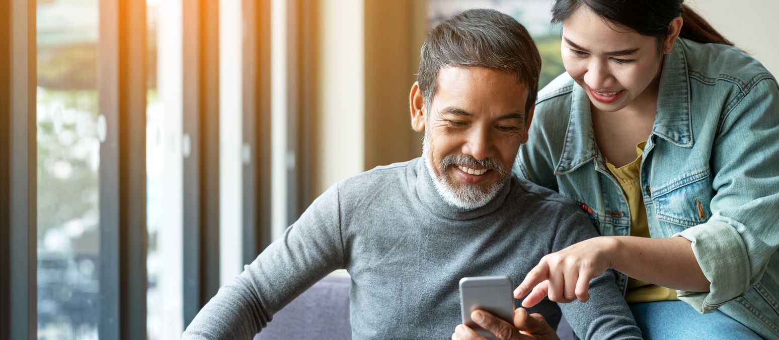 Smile Attractive Stylish Short Beard Mature Asian Man Using Smartphone With Young Woman. Daughter Teach Asian Old Man Or Dad Using Internet Social Media Network Technology With Digital Gadget At Home.