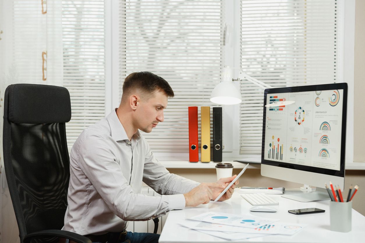 Serious and engrossed business man in shirt sitting at the desk, working at computer with modern monitor, tablet, folders, coffee, documents in light office on window background. Manager or worker.